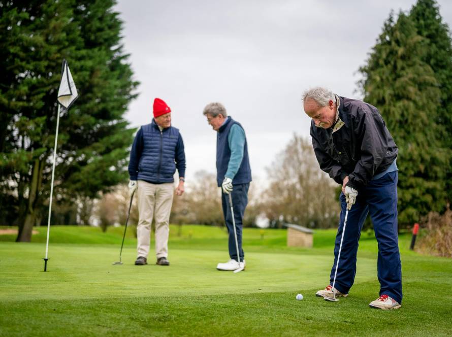 Three Genetlman playing golf. One in foreground putting, two in background talking
